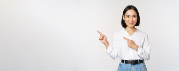 Image d'une jeune femme de bureau souriante, entrepreneur asiatique pointant les doigts vers la gauche, montrant les informations sur le client, carte de bannière de côté sur l'espace de copie, fond blanc - Photo, image