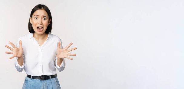 Азиатка смотрит в камеру и кричит в панике. Молодая корейская девушка выглядит встревоженной, паникующей, пожимающей руки и кричащей, стоя на белом фоне - Фото, изображение