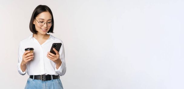 Изображение современной азиатской женщины, смотрящей на мобильный телефон, пьющей кофе на вынос, носящей очки, использующей приложение для смартфонов, стоящей на белом фоне - Фото, изображение