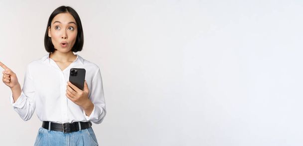 Podekscytowana azjatycka dziewczyna robi zakupy w aplikacji mobilnej, trzyma telefon i wskazuje palcem w lewo na pustej przestrzeni kopii, pokazując tekst promo, stojąc ze smartfonem na białym tle - Zdjęcie, obraz