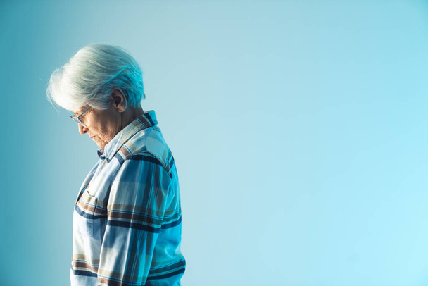 у відставці літня бабуся з сірим волоссям опускає голову в розчарування, вид збоку середній студійний знімок з блакитним тлом
. - Фото, зображення