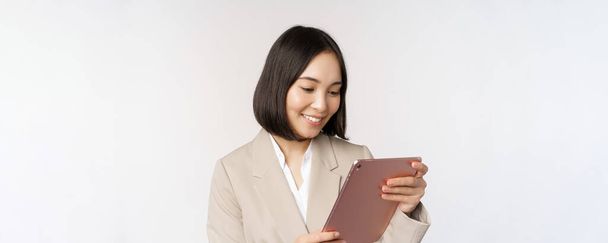 Bild der asiatischen Geschäftsfrau mit digitalem Tablet, Blick auf Gadget und lächelnd, arbeitend, vor weißem Hintergrund stehend - Foto, Bild