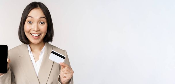 Donna aziendale con volto felice ed entusiasta, che mostra la carta di credito e lo schermo dell'app per smartphone, in piedi in giacca e cravatta su sfondo bianco - Foto, immagini