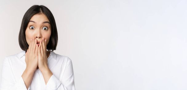 Blisko azjatycka kobieta dysząca, wyglądająca na zszokowaną i pozbawioną mowy, trzymająca się za ręce blisko ust i wpatrująca się w aparat fotograficzny, białe tło - Zdjęcie, obraz