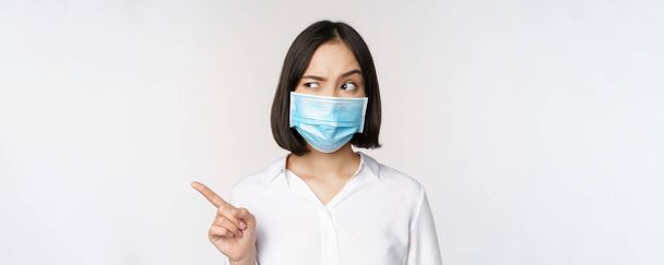 Изображение молодой азиатки в медицинской маске для лица, указывающей влево и смотрящей с подозрительным запутанным выражением лица, стоящей на белом фоне - Фото, изображение
