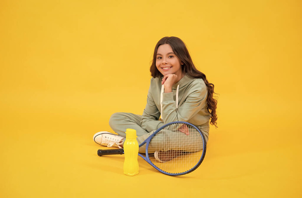 veselé dítě sedět ve sportovním oblečení s badminton raketa a láhev vody na žlutém pozadí, relaxovat - Fotografie, Obrázek