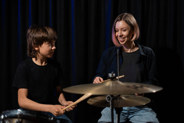 Young caucasian woman teaches a boy to play the drums in the studio on a black background. Music school student - Fotoğraf, Görsel