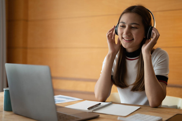 Χαμογελαστή ασιάτισσα μαθήτρια φοράει ασύρματα ακουστικά στο διαδίκτυο, χαρούμενη νεαρή γυναίκα μαθαίνει τη γλώσσα ακούει διάλεξη παρακολουθήσουν webinar γράφουν σημειώσεις ματιά στο φορητό υπολογιστή στο σπίτι, μακρινή εκπαίδευση - Φωτογραφία, εικόνα