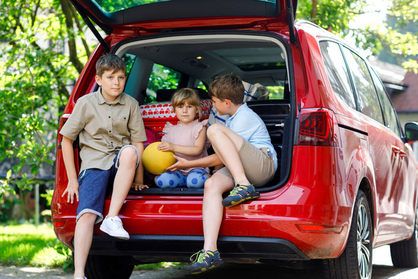 Three children, two boys and preschool girl sitting in car trunk before leaving for summer vacation with parents. Happy kids, siblings, brothers and sister with suitcases and toys going on journey - Fotó, kép