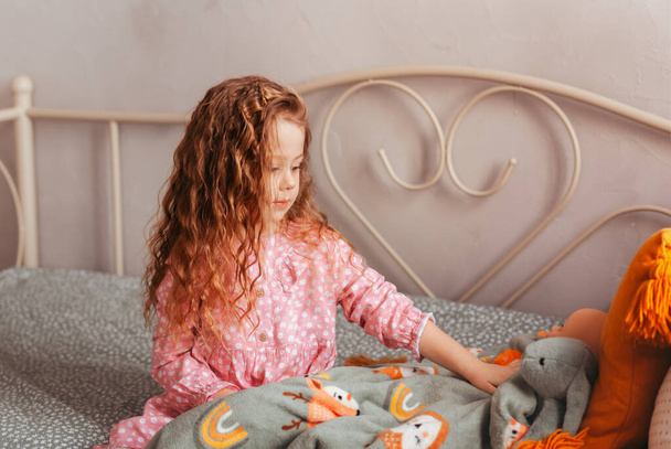 Κορίτσι παιδί πηγαίνει στο κρεβάτι γκρι παιχνίδι λαγουδάκι στην κρεβατοκάμαρά της. Το παιδί ετοιμάζεται να πάει για ύπνο. Ευχήσου γλυκά όνειρα στο αγαπημένο σου παιχνίδι. - Φωτογραφία, εικόνα