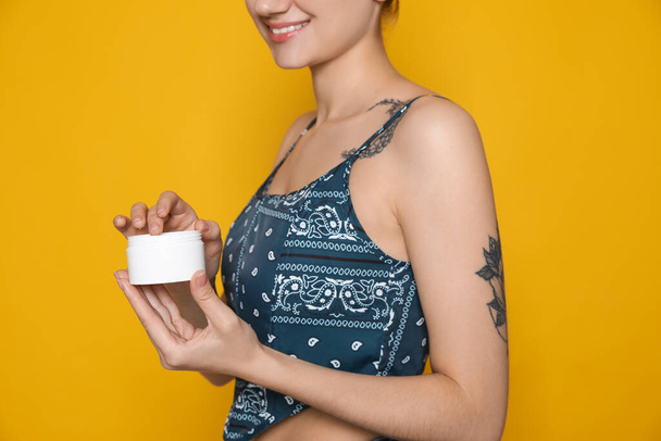 Woman with tattoo holding jar of cream against yellow background, closeup - Foto, afbeelding