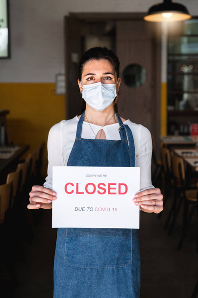 Yüz maskesi ve önlüklü bir kadın işyeri sahibi Covid-19 nedeniyle yazıtların kapalı olduğu bir tabela tutuyor. - Fotoğraf, Görsel