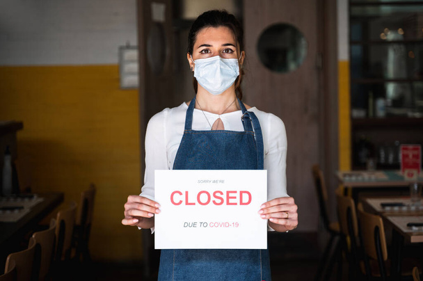 Vertical portrait Female business owner with face mask and apron holding a sign with the inscription closed due to Covid-19 - Foto, Imagem
