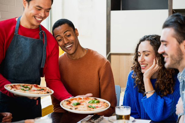 アジアの笑顔の男性とエプロンが彼らに持って来ていることをピザを空腹に見てテーブルに座っている友人の多人種グループ - 写真・画像