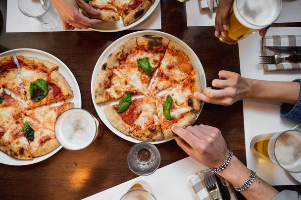 Table de restaurant vue de dessus avec pizza italienne Margherita et verres de bière. Des mains multiraciales méconnaissables saisissant un morceau tranché. - Photo, image
