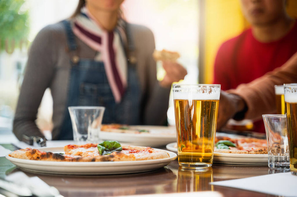 マルゲリータ・ピザのプレートの横にテーブルの上に泡のあるビールのクローズアップグラス。背景にフォーカスしていない人々の認識できないグループは楽しみと食べる. - 写真・画像