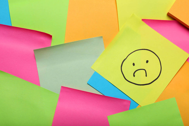 Sticky note with sad face among colorful paper, top view. Space for text - 写真・画像