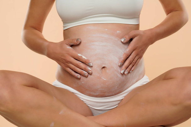 Обрізаний вид вагітної жінки, що наносить косметичний крем на живіт, щоб уникнути розтяжок ізольовані на бежевому
 - Фото, зображення