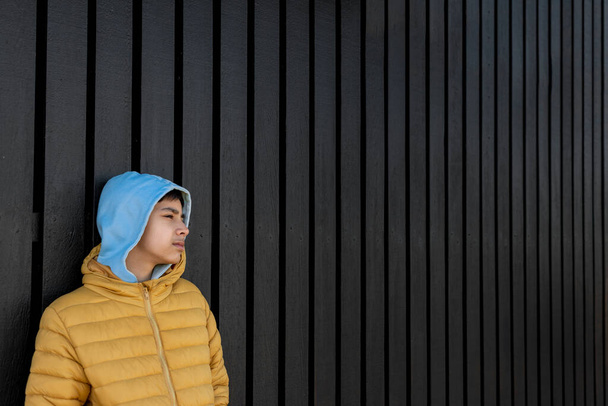 Derékig érő portré a fiatal fiúról, sárga kabátban és kék csuklyában, amint egy fekete fából készült kerítésnél áll, és eltűnik a szülőföldjéről. Háborús gyerekek Ukrajnában. Evakuált gyermekek. - Fotó, kép