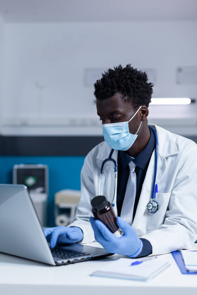 Lekarz specjalista szpitalny trzymający fiolkę z przepisywanym antybiotykiem podczas sprawdzania działań niepożądanych przy użyciu komputera. - Zdjęcie, obraz