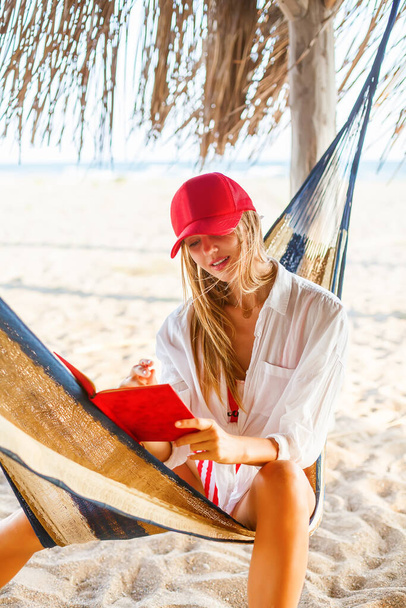 Beautiful woman in red cap writing something in her red planner sitting in a hammock on a beach. Life or job planning in comfortable environment. - Foto, immagini