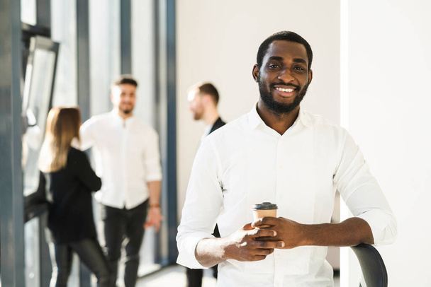 Zwarte mannelijke witte boorden werknemer met een kopje koffie en zijn collega 's op de achtergrond op kantoor - Foto, afbeelding