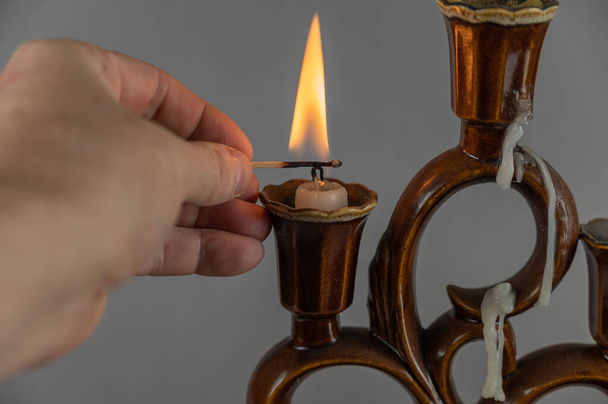 A man's hand lights the candles in the candlestick with a hot match. Brown ceramic candlestick. Gray background. Closeup. Selective focus. - Photo, Image