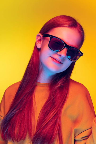 Portrait of trendy young girl posing in stylish black sunglasses isolated over yellow studio background - Foto, Imagen