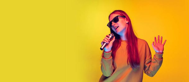 Portrait of young girl singing in microphone, posing isolated over yellow studio background in neon light. Flyer - Photo, image
