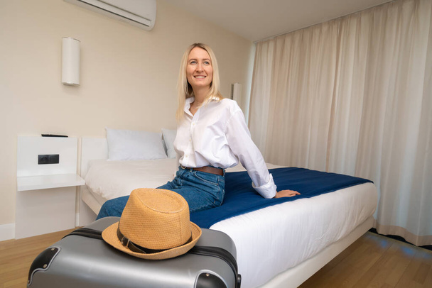 Młoda, uśmiechnięta blondynka w białej koszuli i niebieskich dżinsach siedzi na łóżku w pokoju hotelowym, obok szarej walizki w słomkowym kapeluszu. Pojęcie podróży, turystyki, rekreacji - Zdjęcie, obraz