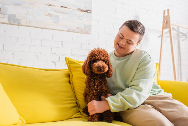 Adolescente alegre con síndrome de Down acariciando caniche en el sofá en casa  - Foto, Imagen
