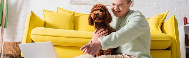 Positive teenager with down syndrome pointing with finger at laptop and hugging poodle at home, banner  - Foto, Imagem