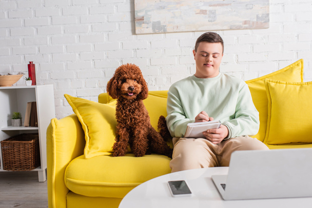 Teenager with down syndrome writing on notebook near devices and poodle at home  - Fotografie, Obrázek