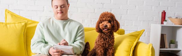 Teen boy with down syndrome writing on notebook near poodle on couch, banner  - Foto, imagen