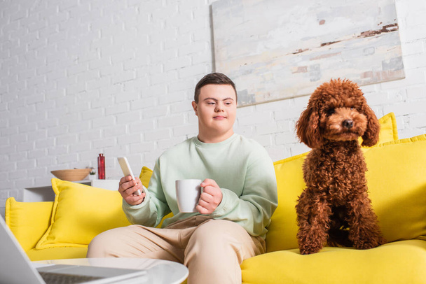 Teenager with down syndrome holding smartphone and cup near poodle on couch  - Фото, изображение
