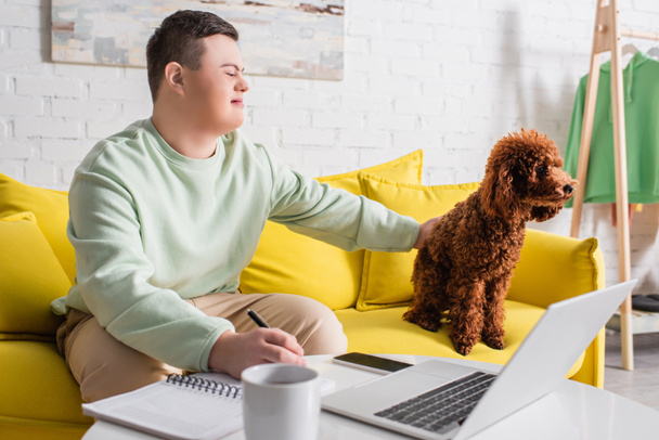 Teenager with down syndrome writing on notebook and petting poodle near gadgets at home  - Fotografie, Obrázek