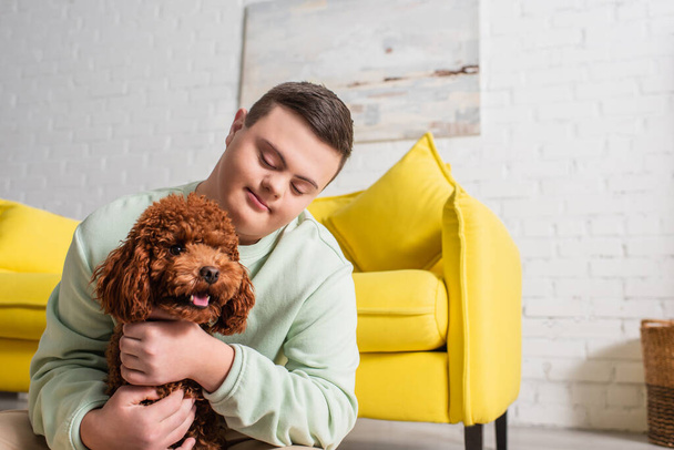 Teen boy with down syndrome hugging brown poodle at home  - Фото, изображение