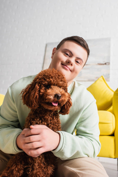 Teen boy with down syndrome hugging poodle and looking at camera in living room  - Фото, изображение