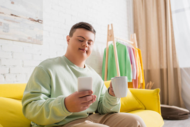 Teenage boy with down syndrome using smartphone and holding cup on couch at home  - Valokuva, kuva