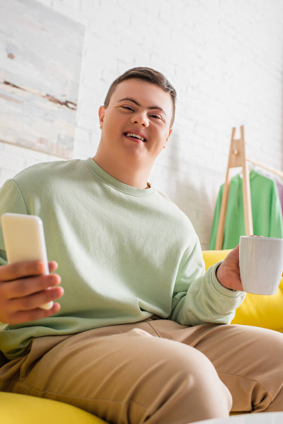 Nízký úhel pohledu veselého teenagera s Downovým syndromem držení mobilu a poháru na gauči  - Fotografie, Obrázek