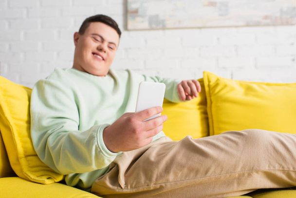 Smartphone en la mano del adolescente borroso con síndrome de Down en el sofá  - Foto, Imagen