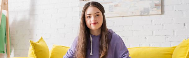Positive teenage girl with down syndrome looking at camera on couch at home, banner  - Фото, изображение