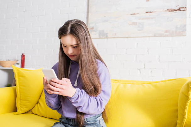 Девочка-подросток с синдромом Дауна использует смартфон на диване дома  - Фото, изображение