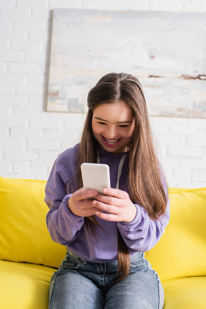 Menina adolescente positiva com síndrome de down usando celular na sala de estar  - Foto, Imagem