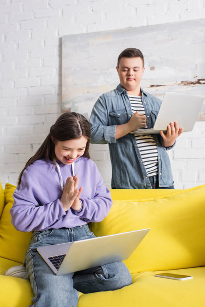 Adolescent joyeux avec le syndrome du duvet en utilisant un ordinateur portable près du smartphone et un ami à la maison  - Photo, image