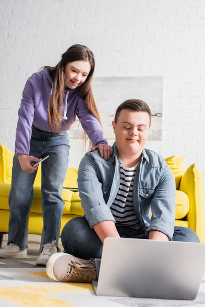 Teenager with down syndrome using laptop near girlfriend with cellphone at home  - Fotó, kép