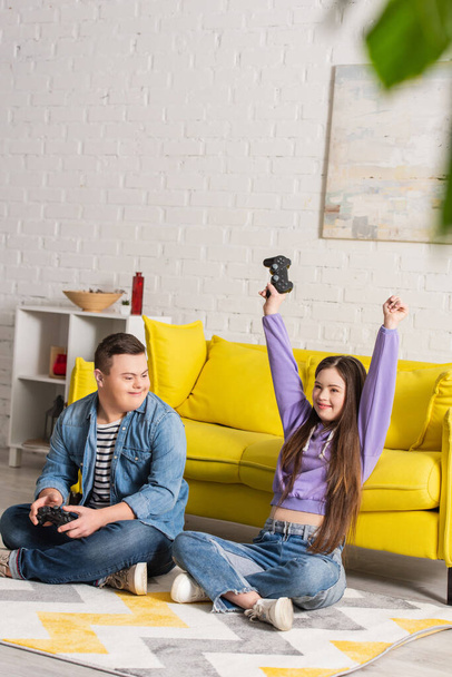 KYIV, UKRAINE - JANUARY 21, 2022: Excited teenager with down syndrome holding joystick near friend at home  - Foto, imagen