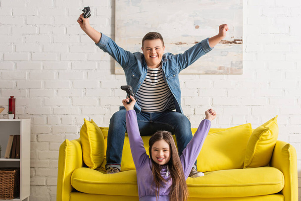 KYIV, UKRAINE - JANUARY 21, 2022: Cheerful teenagers with down syndrome showing yes gesture while playing video game at home  - Foto, immagini