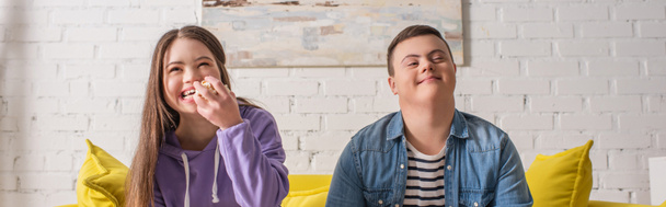 Smiling teenager with down syndrome holding popcorn near friend on couch at home, banner  - Zdjęcie, obraz