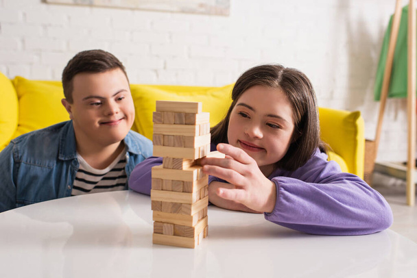 Sorrindo menina adolescente com síndrome de down jogando blocos de madeira jogo perto de amigo em casa  - Foto, Imagem